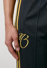 Melni sporta bikses ar dzeltenām sānu svītrām un izšūtu logotipu. Tuvplāns skats, kas parāda auduma tekstūru un šuvju detaļas.