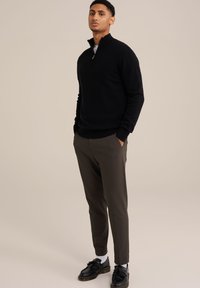 Svart dragkedjetröja i mjukt material, kombinerad med slim-fit olivgröna byxor. Skorna är svarta med tofsdetaljer. Modellen står med händerna i fickorna.