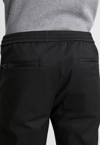 Czarne spodnie o teksturowanej powierzchni z elastycznym pasem na sznurek, dwoma zasuwanymi kieszeniami i dopasowanym krojem, zaprojektowane z myślą o komforcie i funkcjonalności.