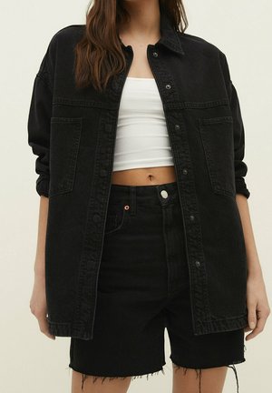Kvinde iført sort oversized denimjakke over hvid cropped tanktop og sorte frynset denimshorts mod en ensfarvet baggrund.