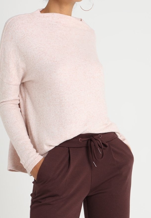 Femme portant un pull à manches longues rose clair et un pantalon à cordon de serrage marron foncé, les mains dans les poches, vue des épaules aux hanches.