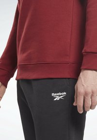 Sweatshirt rouge bordeaux avec poignets côtelés, associé à un pantalon de survêtement noir avec un logo blanc Reebok. Mélange de coton, coupe décontractée, design casual.