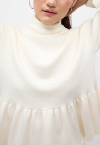 Maglione bianco a maglia con collo alto e orlo peplum. Il tessuto presenta una finitura texture, con sottili variazioni di colore e struttura.