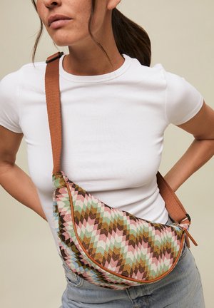 Mujer con camiseta blanca y vaqueros, con bolso bandolera de tela con estampado en zigzag en tonos rosa, verde, azul y marrón.