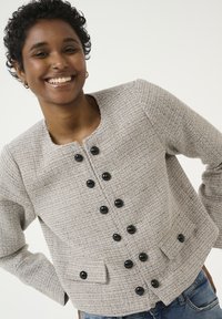 Texturált, bézs tweed dzseki kerek nyakkal, nyolc fekete gombbal elöl, és két elülső zsebbel.