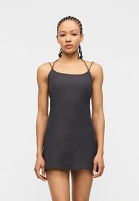 Γυναίκα με πλεγμένα μαλλιά που φορά ένα αμάνικο μαύρο μίνι φόρεμα με λεπτές τιράντες με ξύλινα χάντρες, σε απλό ανοιχτό φόντο.