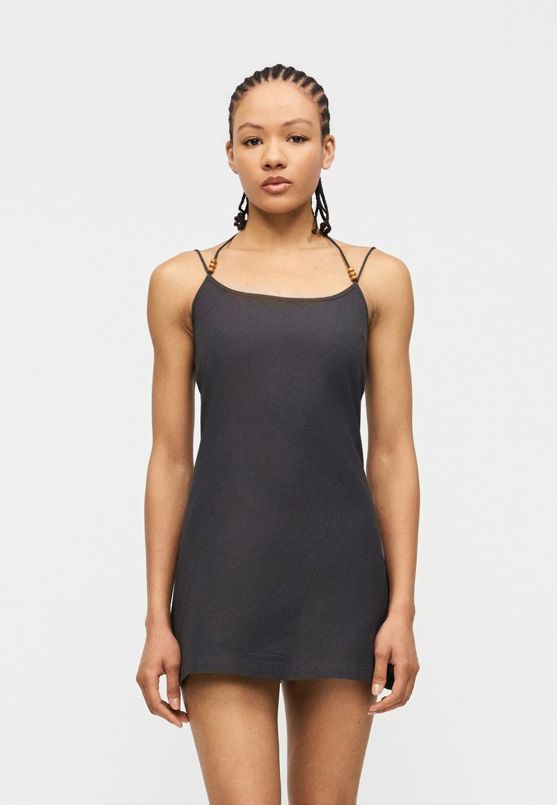 Γυναίκα με πλεγμένα μαλλιά που φορά ένα αμάνικο μαύρο μίνι φόρεμα με λεπτές τιράντες με ξύλινα χάντρες, σε απλό ανοιχτό φόντο.