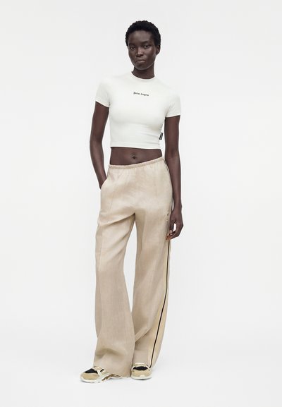 Λεπτή όρθια γυναίκα με κοντά μαλλιά που φοράει λευκό cropped τοπ με κοντά μανίκια, μπεζ παντελόνι με φαρδιά πόδια και ρίγα στο πλάι και μπεζ sneakers.