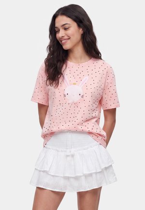 Frau trägt ein pinkes Shirt mit kleinen schwarzen Herzen und einem Hasengesicht mit Krone, kombiniert mit einem weißen, gerafften Rock.