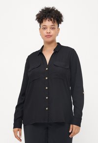 Chemise noire à manches longues en tissu léger avec un devant boutonné, deux poches poitrine et des boutons dorés. Texture lisse, coupe décontractée.