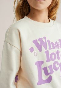 Mikina krémovej farby z jemnej látky, s veľkým fialovým nápisom a dekoratívnymi prvkami v podobe kvetinových a hviezdičkových tvarov.