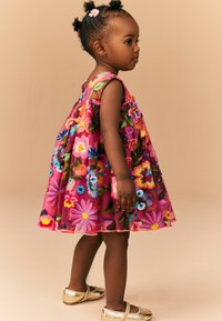 Rosa broderad klänning med färgglada blommönster, ärmlös design, lagerkjol och gula skor. Enkla håraccessoarer fulländar looken.