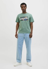 Zelené bavlněné tričko s grafickým designem s textem "Jack & Jones". Spárováno se světle modrými džíny a bílými teniskami.