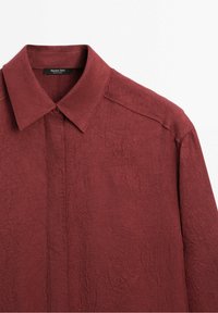 Chemise bordeaux à manches longues avec un grand col pointu et un tissu subtilement texturé, présentée sur fond blanc.