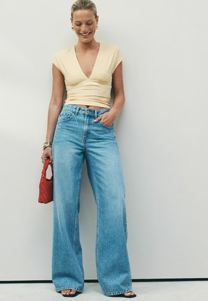 Frau trägt ein beiges Kurzarmtop und weite blaue Jeans, hält eine kleine rote gewebte Tasche und lächelt vor einem schlichten Hintergrund.