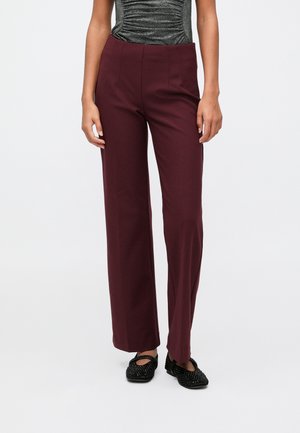 Mulher vestindo calças burdeos de cintura alta e perna reta, com um top drapeado cinza metálico e sapatos planos pretos texturizados.