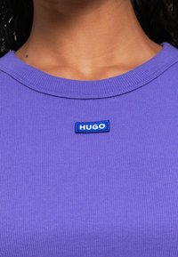 Primo piano di un collo di una maglietta rigata viola con una piccola toppa rettangolare blu etichettata "HUGO" centrata sotto il colletto.