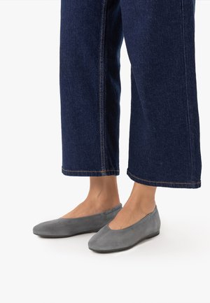 Ben iført mørkeblå brede jeans og grå ruskindssko med flad hæl på hvid baggrund.
