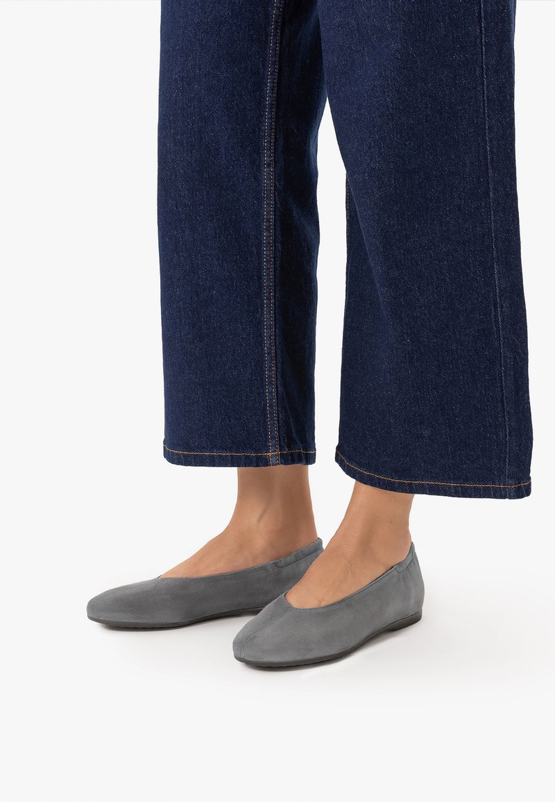 Jambes portant un jean large bleu foncé et des chaussures plates en daim gris sur un fond blanc.