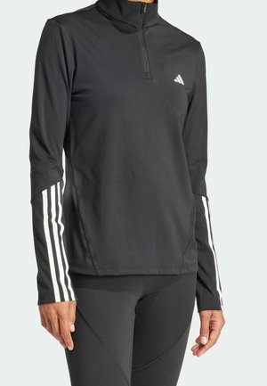 Haut de sport noir à manches longues avec fermeture éclair partielle, détails blancs à trois bandes sur les manches et petit logo blanc sur la poitrine gauche, porté avec des leggings noirs.