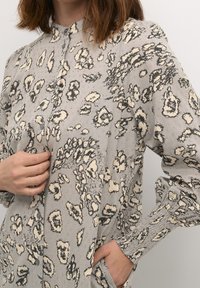 Grå knäppblus med ett leopardmönster i beige och svart, med ståkrage och rynkade manschetter.