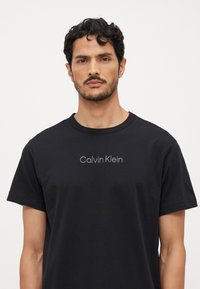 Crna pamučna majica s okruglim izrezom i kratkim rukavima. Istaknuto „Calvin Klein“ brendiranje u sivoj boji preko prsnog dijela.