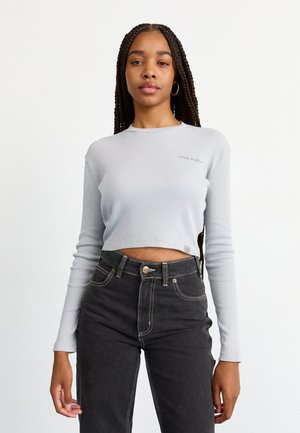 Giovane donna con lunghi capelli intrecciati, che indossa un top a maniche lunghe a coste grigio chiaro, cropped, e jeans neri a vita alta, su sfondo bianco.