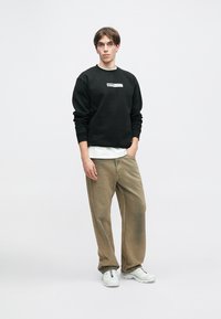 Černá mikina s bílým logem, vrstvená přes bílou košili, doplněná volnými béžovými kalhotami a bílými teniskami. Jednoduchý, ležérní design.