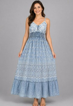 Femme souriante portant une robe maxi bleue sans manches avec des motifs floraux et en dentelle, debout devant un fond gris uni.