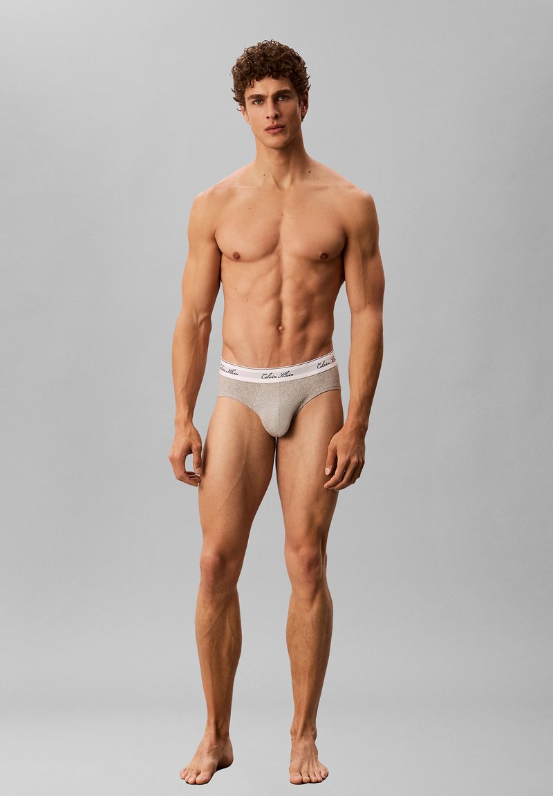 Jeune homme musclé aux cheveux bouclés, debout pieds nus sur un fond uni, portant un slip beige Calvin Klein.