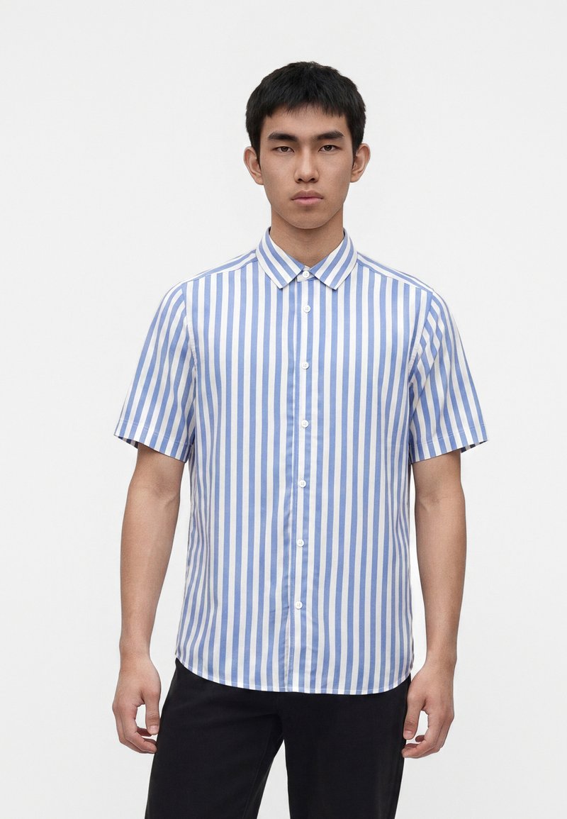 Jeune homme portant une chemise boutonnée à manches courtes à rayures verticales bleues et blanches et un pantalon noir, debout devant un fond blanc uni.