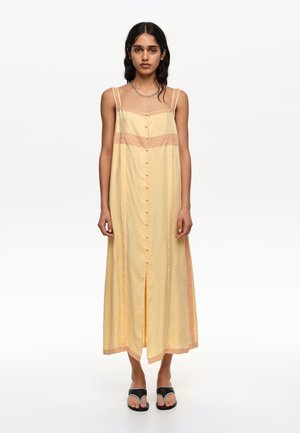 Mujer joven con cabello largo y oscuro, vestida con un vestido largo sin mangas de color amarillo pálido con botones frontales y sandalias negras de punta abierta sobre fondo blanco.