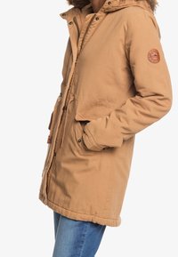Giacca parka beige con cappuccio, chiusure a zip e a bottoni, girovita con coulisse e una patch rotonda con logo sulla manica sinistra. Tessuto strutturato.