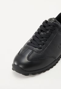 Black athletic shoe featuring a mix of smooth and textured leather, round toe, thick rubber sole, and flat black laces.