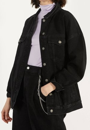 Personne portant une veste en jean noire avec des boutons argentés, un pantalon noir avec un détail chaîne, et un haut col roulé violet clair.