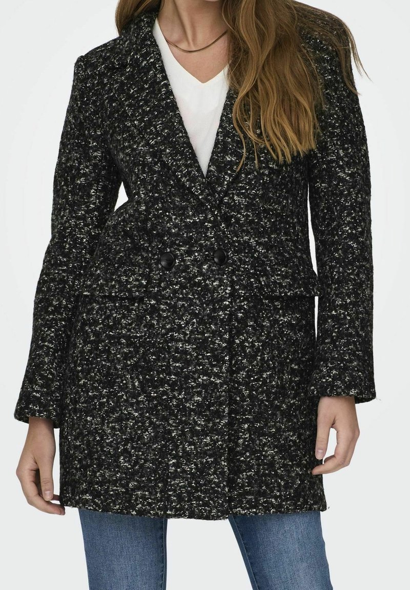 Femme portant un manteau croisé texturé noir et blanc sur un haut blanc et un jean bleu, avec des cheveux longs et lâches.