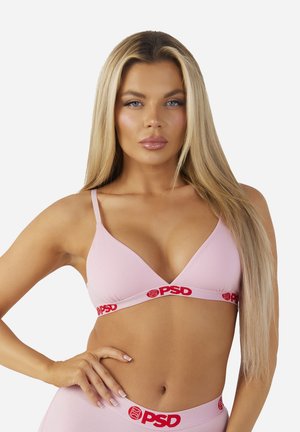 Frau mit langen blonden Haaren, die einen hellrosa Bralette und passende Unterwäsche mit rotem PSD-Logo auf den elastischen Bändern trägt.