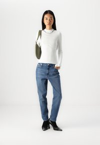 Balta ribota adīta blūze ar augstu apkakli, zila džinsu bikses, melni norādīti papēžu apavi ar spīdīgu purngalu un zaļa plecu soma.