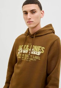 Brun hoodie med snøre, med en stor gulgrøn grafisk tekst der står "JACK & JONES Denim Goods" på forsiden. Blød tekstur.