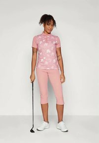 Ροζ πόλο μπλούζα με λουλουδάτο σχέδιο και κοντά μανίκια, συνδυασμένη με ανοιχτό ροζ καπιρί παντελόνι. Το μοντέλο κρατάει ένα γκολφ, φορώντας λευκά αθλητικά παπούτσια.