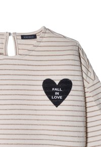 Chemise crème et or rayée avec un col rond, une ouverture en forme de clé au dos, et un patch en forme de cœur noir indiquant "FALL IN LOVE".