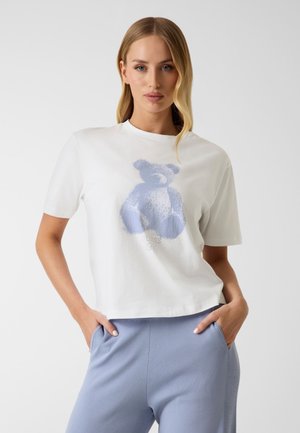 Femme blonde debout, les mains dans les poches, portant un t-shirt blanc avec un motif d’ours en peluche bleu et un pantalon bleu clair.