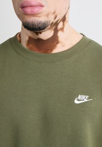 Olivno zelena bombažna majica s krožnim izrezom in belim Nike logotipom na prsih. Gladka tekstura, minimalističen dizajn.