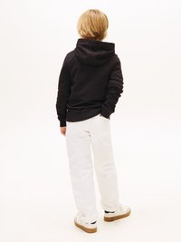 Melnā kapuce ar kapuci, baltas brīvas bikses un baltas sporta apavi. Mati ir vidēja garuma un gaišas krāsas. Fons ir vienkāršs balts.