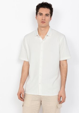 Młody mężczyzna ubrany w białą koszulę na krótki rękaw z guzikami oraz beżowe spodnie typu cargo, stojący na jednolitym białym tle.