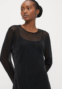 Μαύρο φόρεμα με μακριά μανίκια και διάφανο, λαμπερό κάλυμμα, με στρογγυλή λαιμόκοψη και εφαρμοστή σιλουέτα, που αναδεικνύει διακριτική λάμψη.