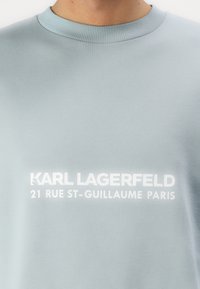Svetlo modra trenirka z rebrastim ovratnikom in belim tiskanim besedilom: "KARL LAGERFELD 21 RUE ST-GUILLAUME PARIS."