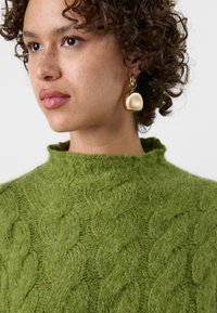 Pull en tricot vert avec un col montant et un motif de torsade texturé. Boucles d'oreilles pendantes dorées de forme arrondie complètent le design.
