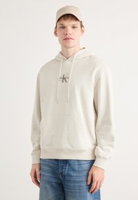 Beige Kapuzenpullover mit Fronttasche und Kordelzug, ausgestattet mit einem kleinen Calvin Klein Logo in Braun. Kombiniert mit hellblauen Jeans.