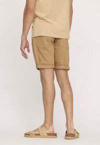 Shorts en coton beige avec des revers, présentant des poches arrière. Portés avec des sandales beiges. L'arrière-plan est d'une couleur claire unie.